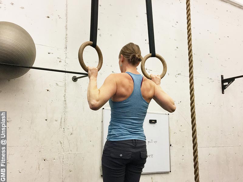 A woman performing a pull-up hold on Olympic rings with a climbing rope in the background, symbolising strength and fitness challenges.