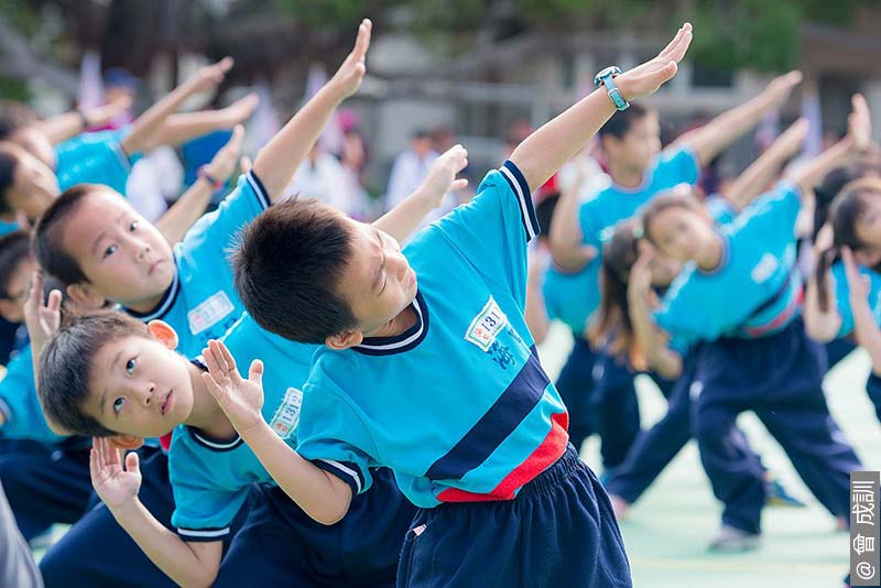 A group of young students in blue sports uniforms engaging in Radio calisthenics at Haikou Elementary School's Sports Day in Zhunan. The children are stretching their arms up and to the side, showing concentration and coordination in their movements.