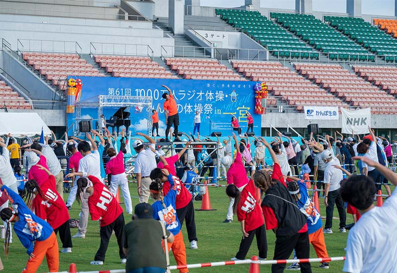 A wide-angle photograph of a large Radio-Taiso gathering in a stadium, with participants of various ages performing synchronised exercises. In the background, banners with Japanese writing and logos of NHK and Japan Post are visible, underlining the event's significance and support.