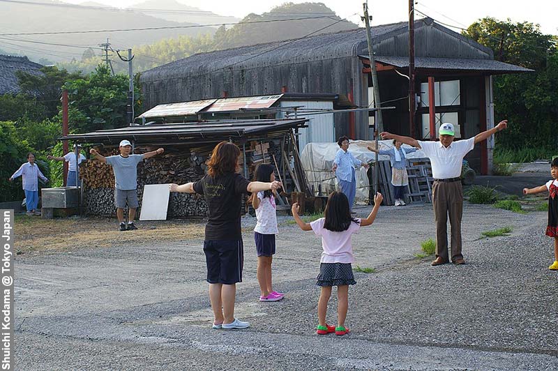 Residents of varying ages perform Radio calisthenics in a rural area, with the backdrop of modest buildings and stacked firewood, under the soft glow of morning light.