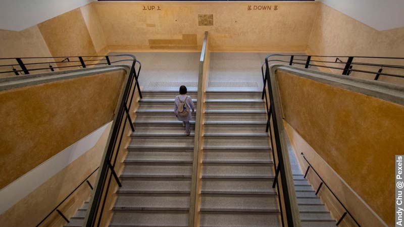 A photorealistic image of a woman with a backpack, actively walking up the stairs in an office building, with the aim of overcoming physical activity barriers. Symbolising the choice of incorporating physical activity into a daily routine, over more sedentary options like elevators.