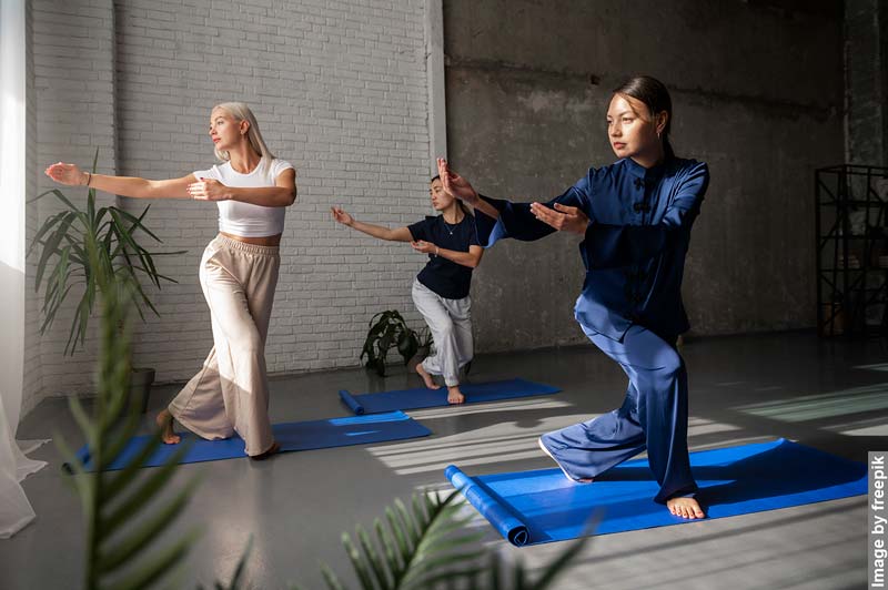 Three women of different ages practicing Tai Chi in a well-lit studio with plants and yoga mats, focusing on their movements and balance, showcasing the benefits of Tai Chi for physical and mental health.