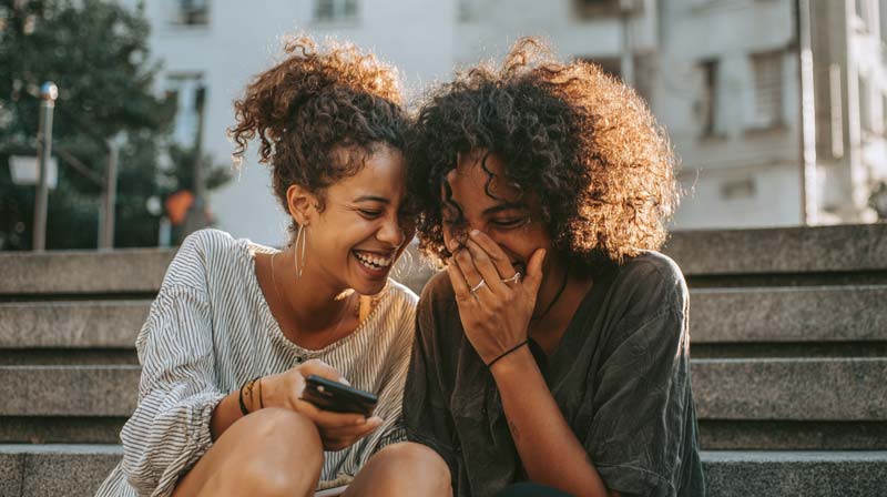 Two young women sit close together on outdoor steps, leaning towards each other and laughing over something on a mobile phone, one resting a hand on the other’s shoulder while the other covers her mouth as soft afternoon light highlights their faces and easy friendship.