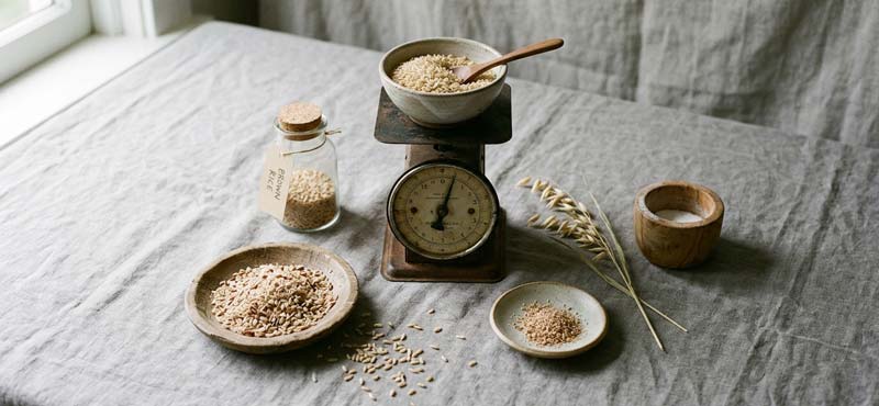 Wooden bowl of brown rice resting on a vintage kitchen scale, with scattered grains, a glass jar, and a small dish on a grey linen surface.