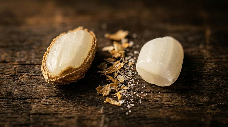Macro image of a split rice grain showing one half with bran intact and the other polished white, with fine rice fragments on a dark wooden surface.