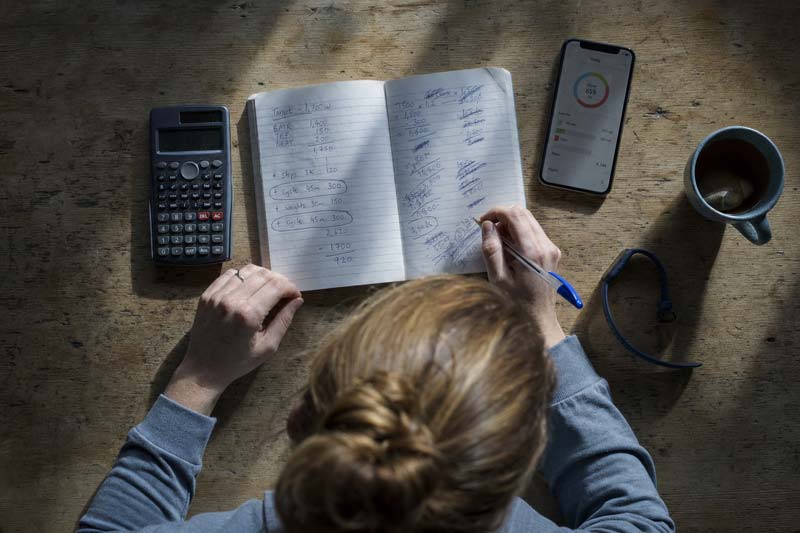 Overhead view of a woman checking handwritten calorie calculations beside a calculator, phone and fitness tracker while considering whether to eat back exercise calories.