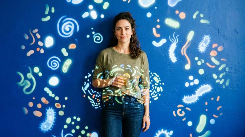 A woman holds a drink while projected microbial shapes gather across the background around her abdomen.