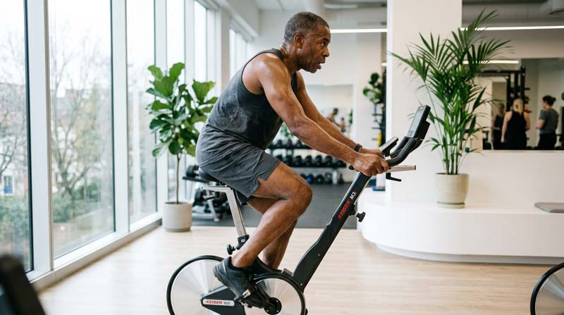 An older man exercises on an upright stationary bike with focused effort and controlled breathing in a bright modern training space.