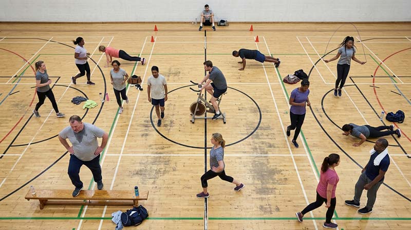 A group of ordinary adults perform different interval exercises and recovery movements across a marked community sports hall floor, viewed from above.