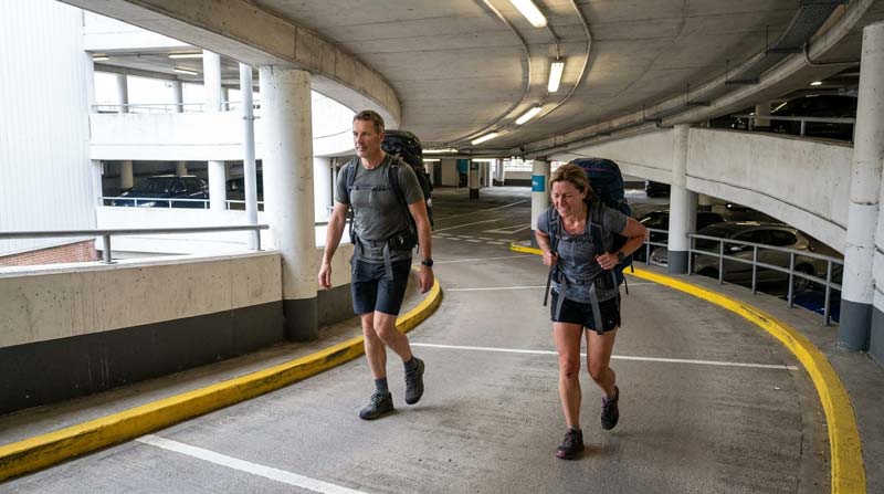 Two adults rucking up a curved car park ramp with similar backpacks while one maintains a steady stride and the other shows greater physical effort.