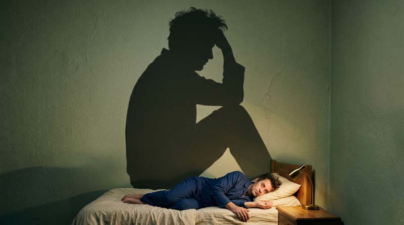 A tired man lies curled in bed while his oversized shadow sits upright on the wall, holding its forehead.