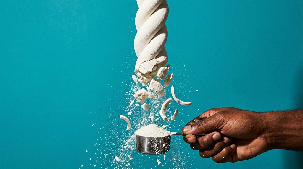 A dark-skinned hand holds a steel scoop beneath a crumbling white helix and falling powder, symbolising collagen peptides.