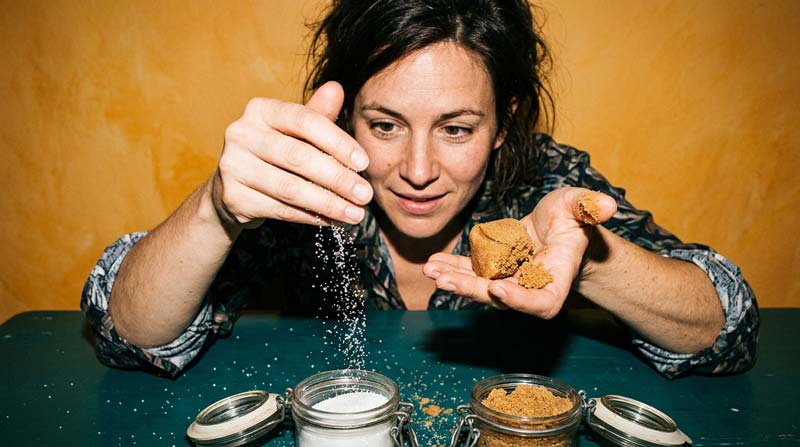 A woman compares brown and white sugar by touch, with white grains falling through her fingers and brown sugar holding together in her other palm.