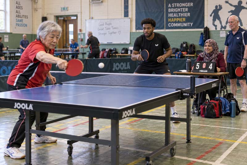 Older woman playing table tennis with a younger man at a community club, accompanying an article on ageing and health.