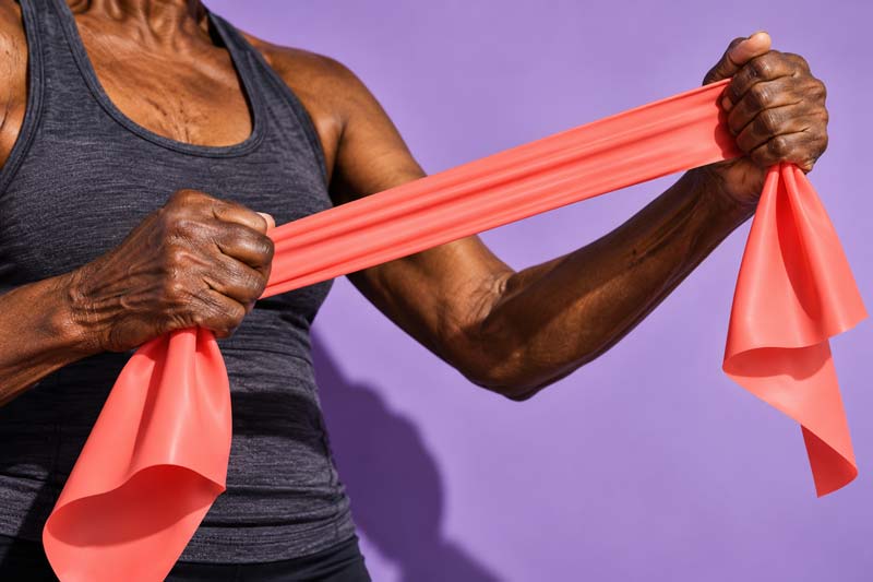 Close-up of an older woman’s hands stretching a coral resistance band against a lilac background.