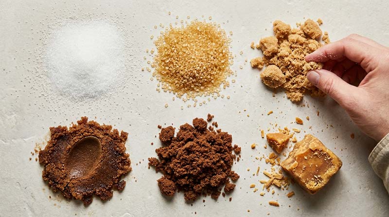 Six samples of brown and white sugar, including demerara, muscovado and a broken jaggery block, display contrasting textures on pale stone.