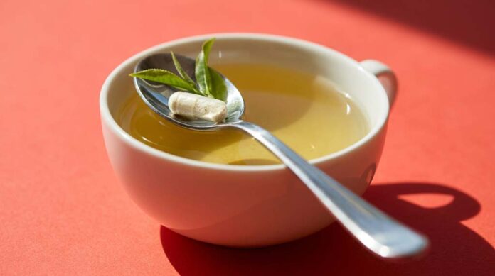 A capsule of L-theanine and a fresh tea shoot rest in a reflective teaspoon across a porcelain cup of tea.