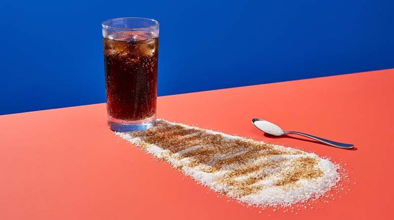 A glass of cola casts an oversized shadow made from sugar granules, with a small spoonful beside it on a coral surface.