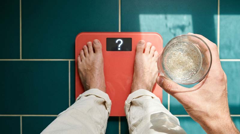Bare feet rest on a coral bathroom scale displaying a question mark, while a hand holds a glass of cloudy drink above teal floor tiles.