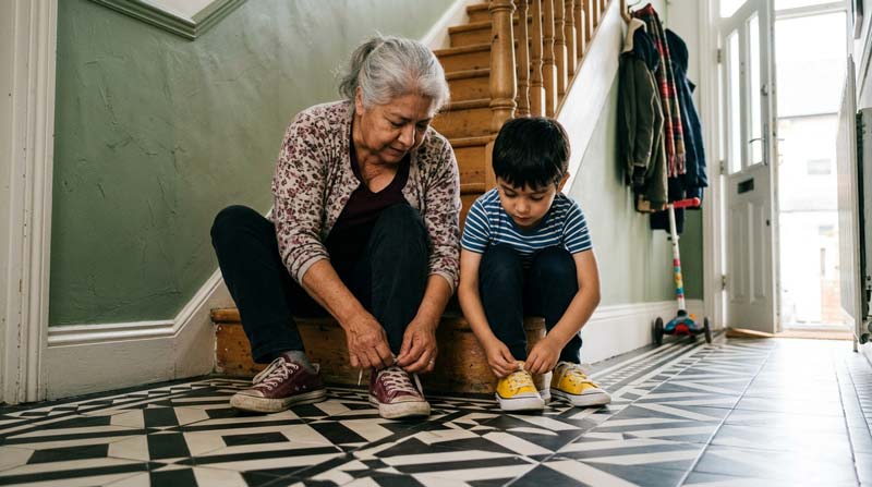 An older woman and a young boy sit beside each other on a hallway stair, tying their trainer laces.