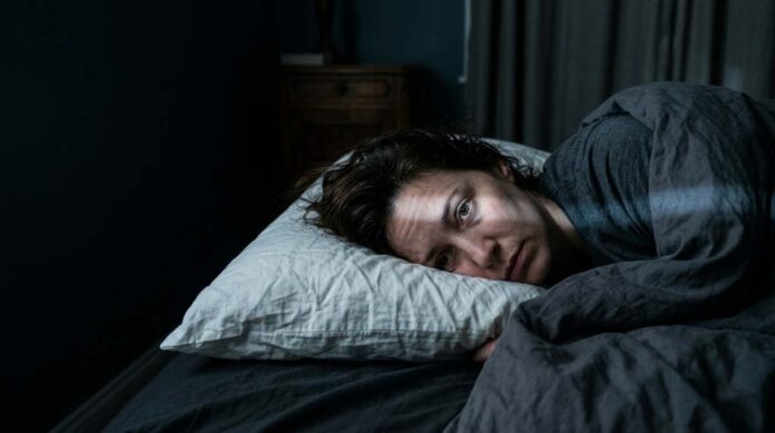 A woman who is tired but can't sleep lies on her side, a narrow strip of light illuminating her open eye.
